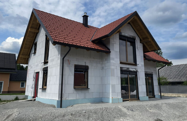 Dvonadstropna hiša v gradnji z belimi zunanjimi stenami, velikimi okni in rdečo streho iz opeke. Zgrajena je iz masivne gradnje in ponuja varjeno hišo. Dvorišče je nedokončano, stavbo obdaja gramoz. Nebo je delno oblačno.